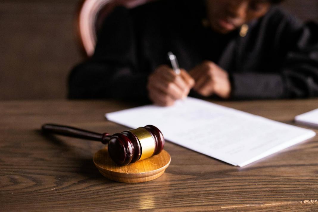 Ciocan de judecător pe masă în sala de judecată, cu un judecător scriind documente în fundal.